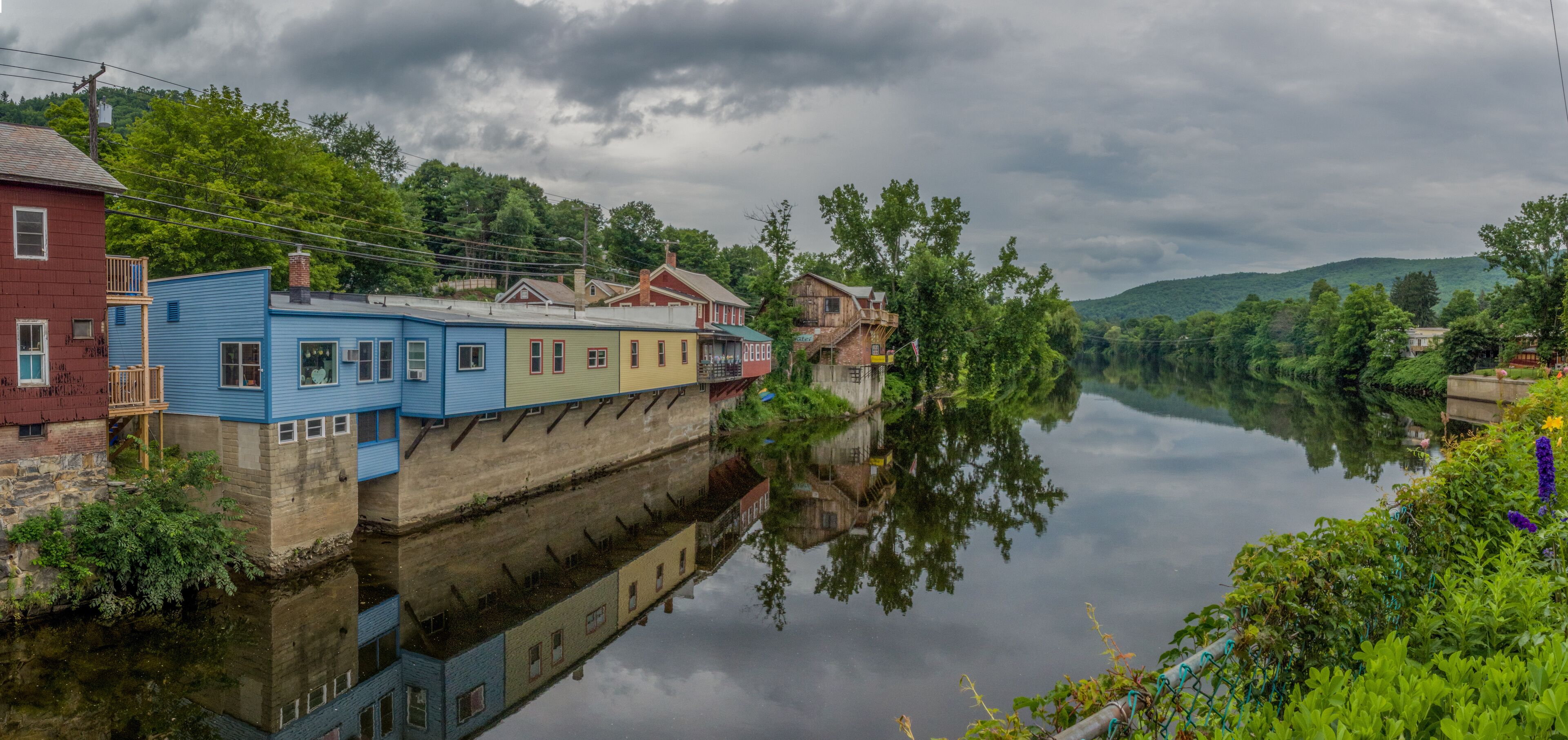 A river with buildings reflected on the water, surrounded by greenery and hills.