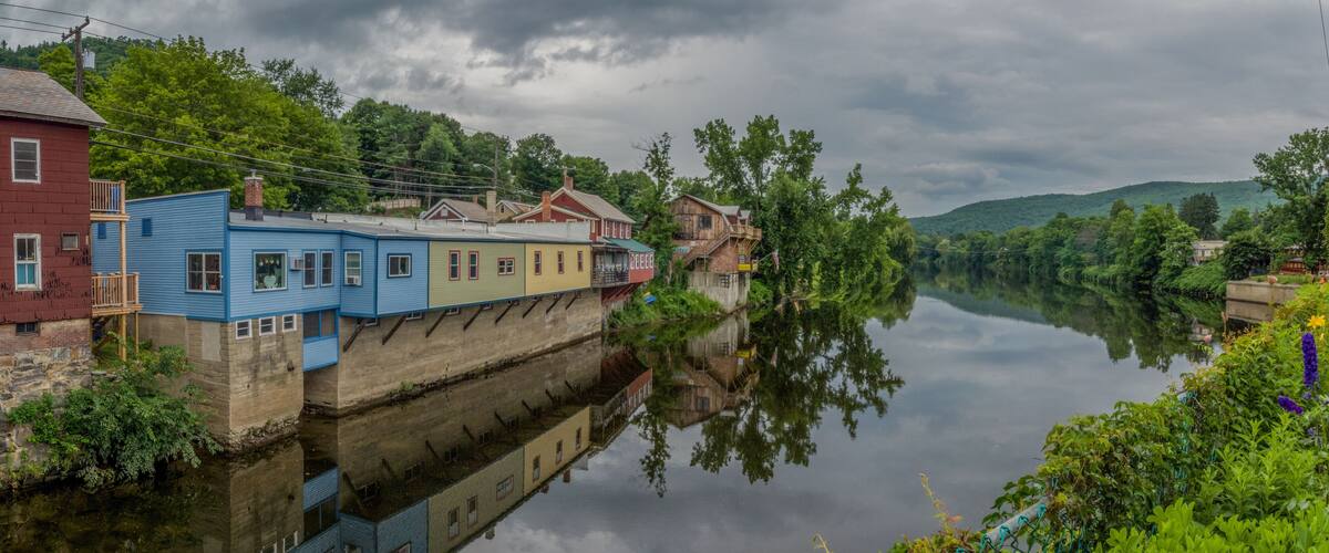 A river with buildings reflected on the water, surrounded by greenery and hills.