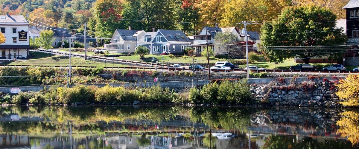 A lakeside town with autumn-colored trees, a stone bridge, and houses reflecting in the water.