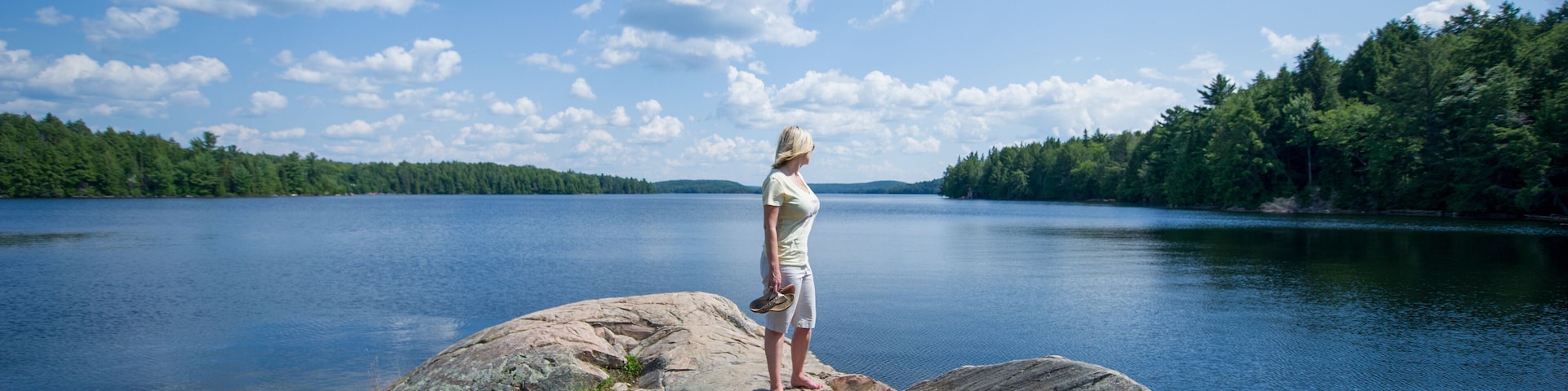 Young woman standing on a rock looking at Smoke lake in Algonquin Provincial Park, Muskoka Ontario Canada. The blue sky is showcasing beatiful fluffy clouds