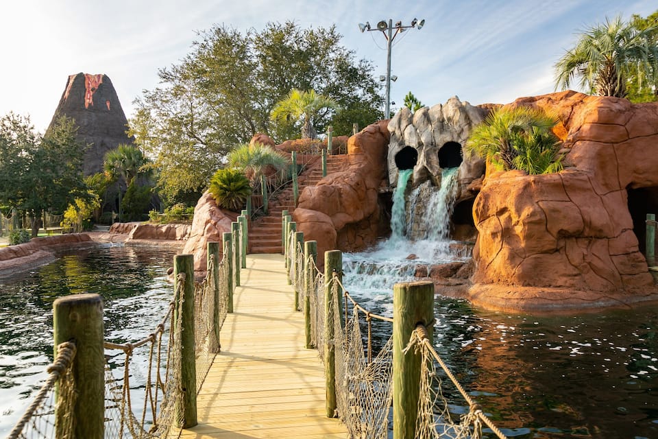 A wooden boardwalk leading to a waterfall, surrounded by rocky formations and palm trees.