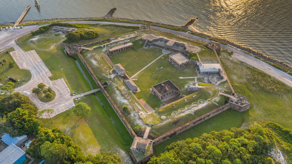 An aerial view of a historic fort with a moat, surrounded by greenery and a body of water.