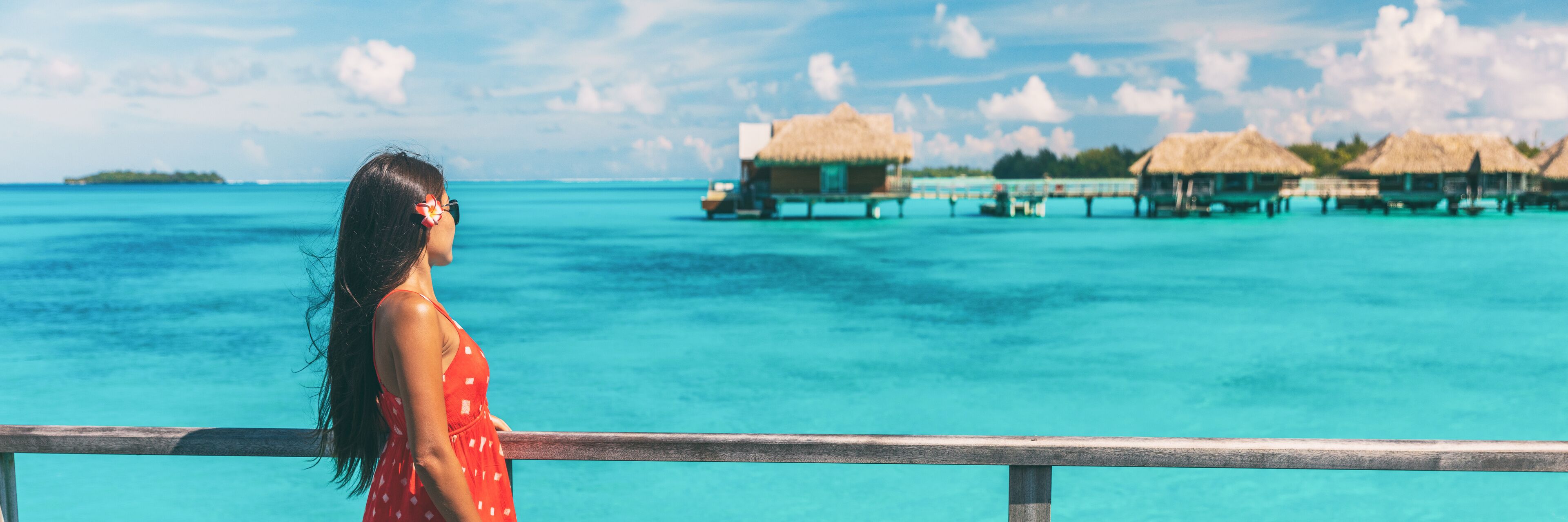 A woman in a red dress overlooking turquoise ocean waters with overwater bungalows.