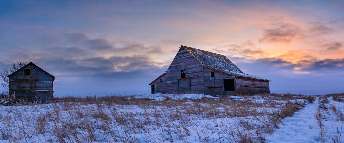 Two wooden barns on a snowy hill with a sunset sky.