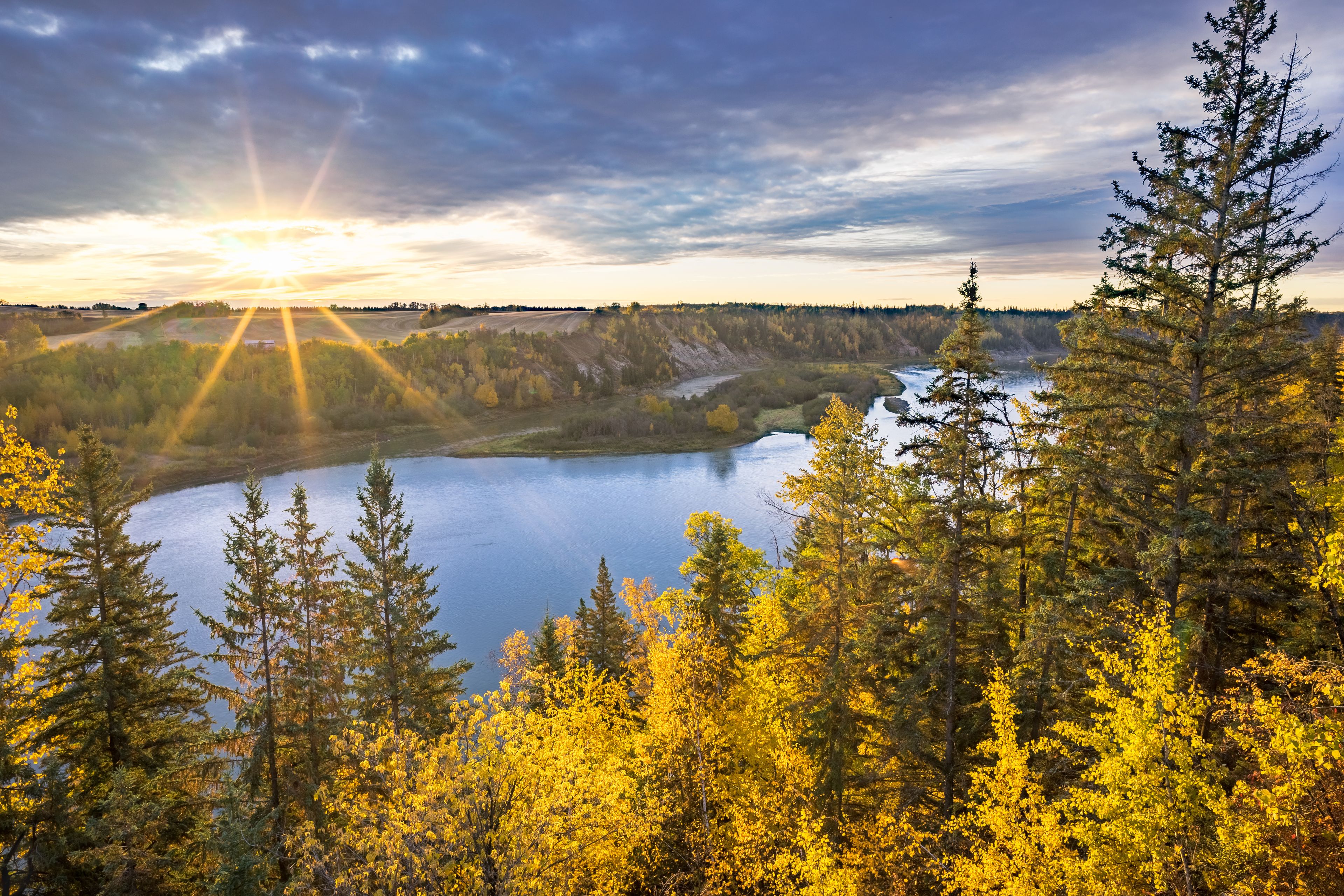 A river winding through a forest with autumn foliage, with the sun setting in the background.