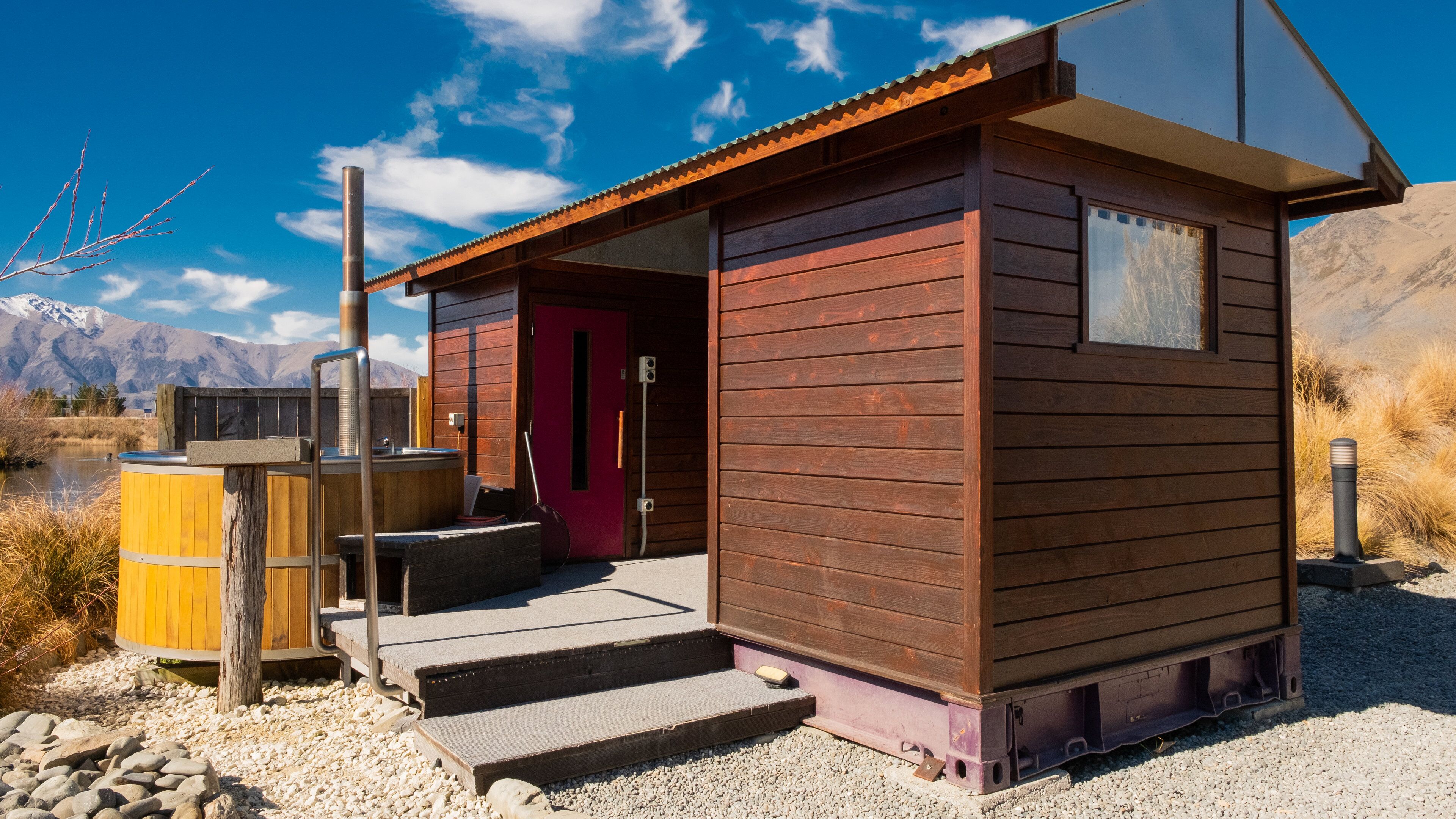 A wooden cabin with a red door, a hot tub, and steps leading up to it, set against a mountainous backdrop.