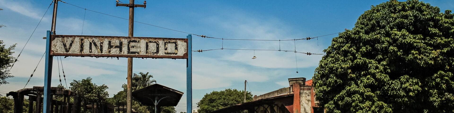 A railway station with a sign reading 'VINHEDO,' surrounded by greenery and a clear blue sky.