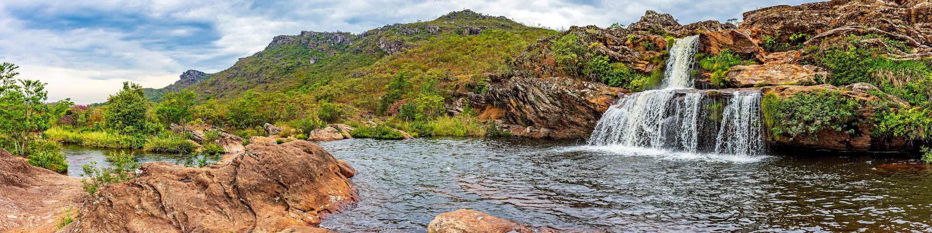 A waterfall cascading over rocks into a river surrounded by lush greenery and hills.