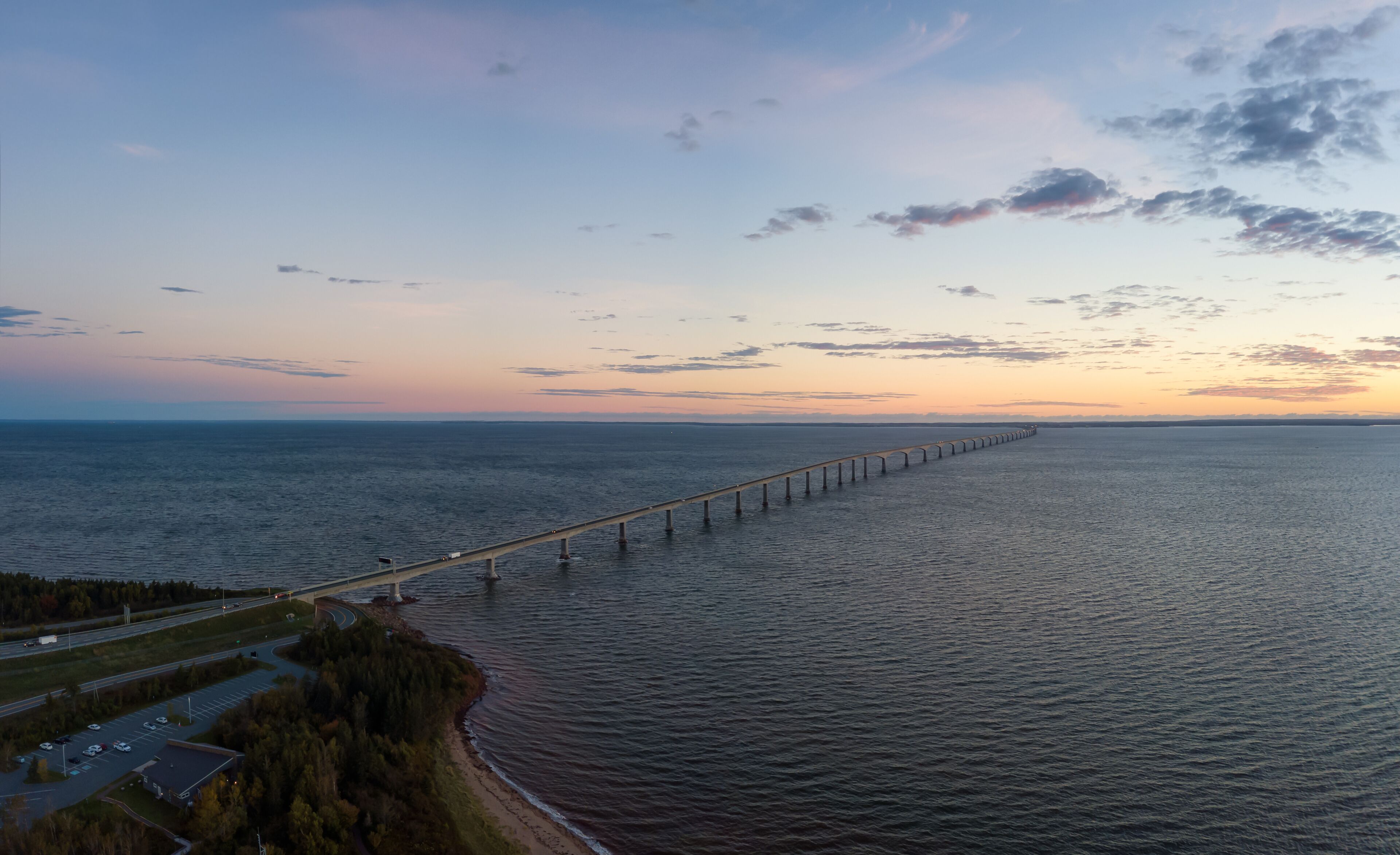 A coastal road with a long bridge extending into the sea, surrounded by trees and a clear sky.