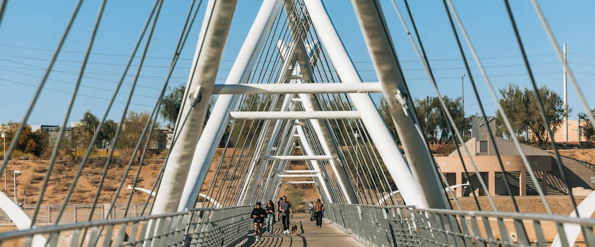 A pedestrian bridge with metal railings and cables, surrounded by dry vegetation and a clear sky.