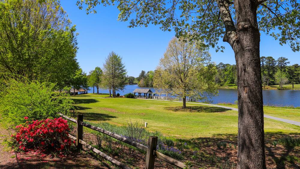 A gorgeous summer landscape in the garden with a lake and lush green trees, grass and plants, a brown wooden boathouse, red flowers, and wooden fence with blue sky at Callaway Gardens in Pine Mountain