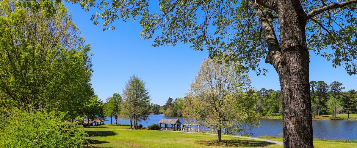 A lakeside park with a wooden fence, red flowers, and a gazebo by the water.