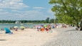 Ein voller Strand mit Menschen, Sonnenschirmen und einem See im Hintergrund.