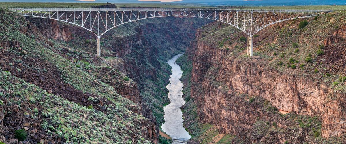 A bridge over a canyon with a river below, surrounded by mountains and a sunset sky.