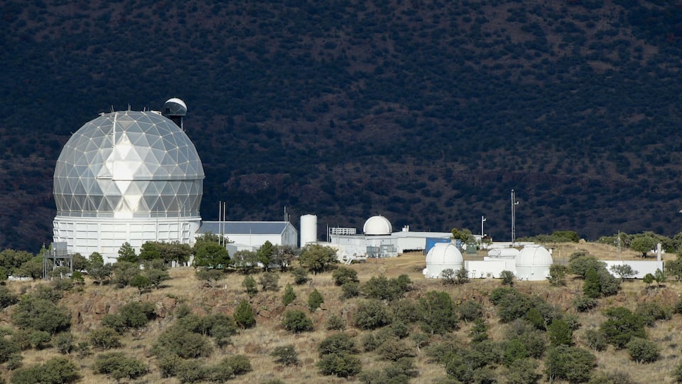 A large observatory dome on a hill with surrounding trees and a mountainous backdrop.