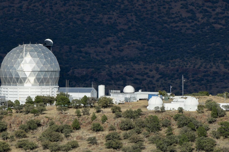 Una gran cúpula de observatorio en una colina, rodeada de árboles y con montañas al fondo.