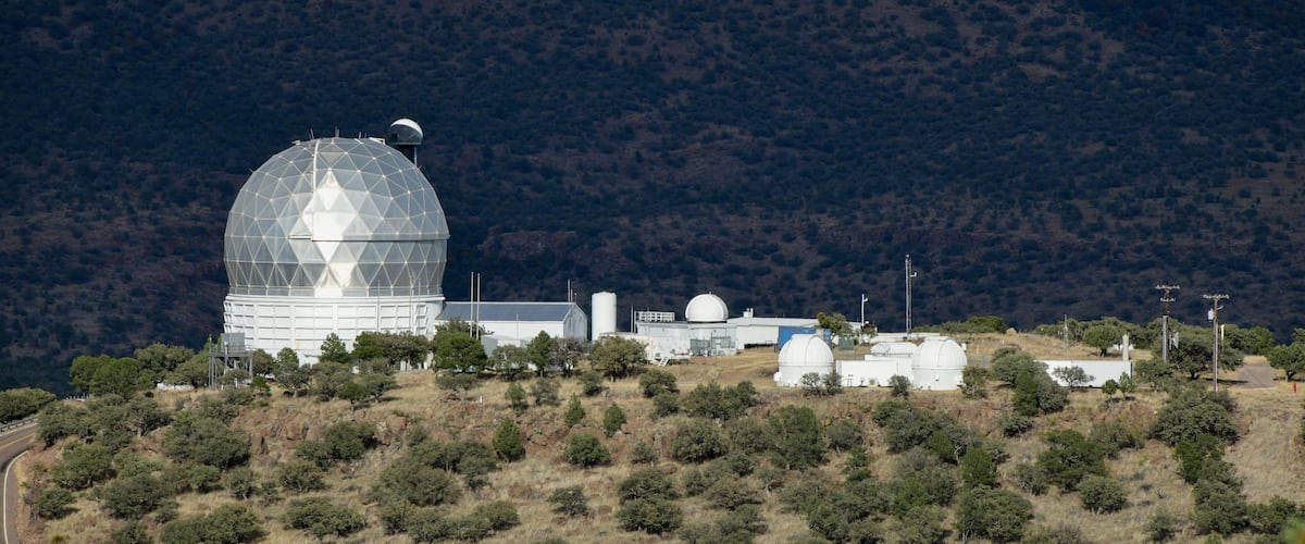 A large observatory dome on a hill with surrounding trees and a mountainous backdrop.