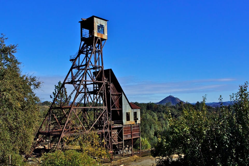 A rusted metal tower with a wooden house at its base, surrounded by dense greenery and a mountain in the background.