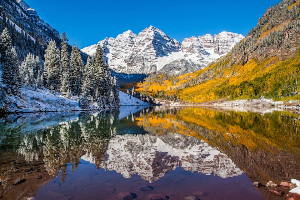 Um lago montanhoso com picos nevados, refletindo as montanhas, e árvores com cores de outono.