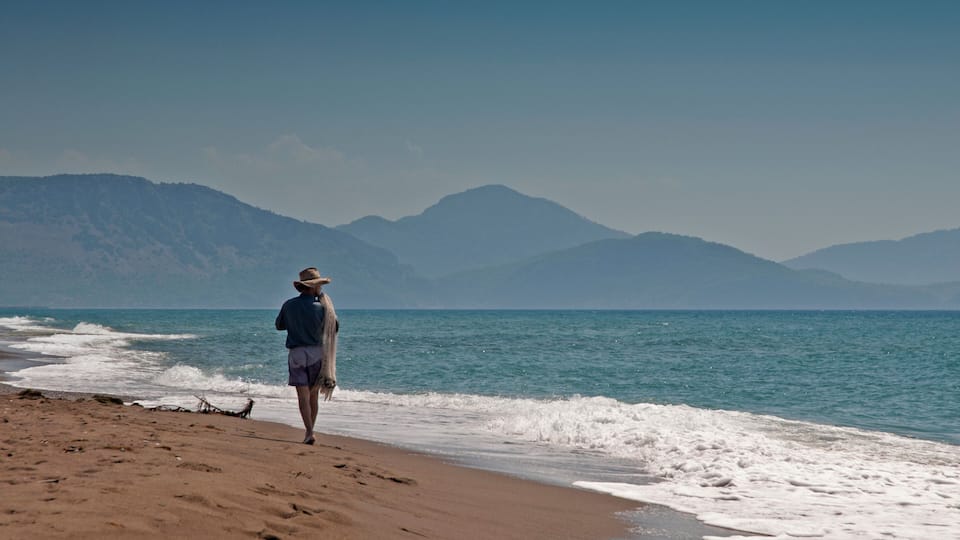 A person walking along a sandy beach with waves crashing onto the shore, and mountains in the background.