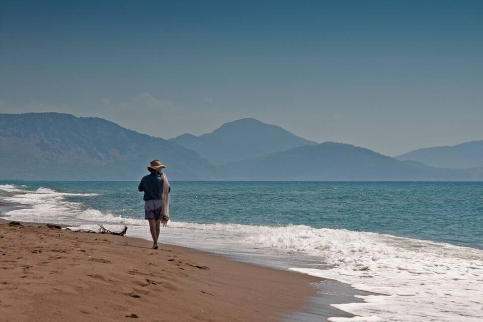 A person walking along a sandy beach with waves crashing onto the shore, and mountains in the background.