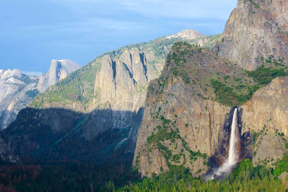 Una cascada que es precipita per un penya-segat empinat, envoltada de bosc dens i muntanyes.