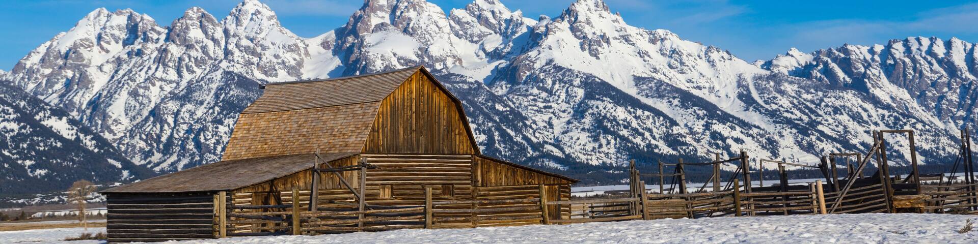 A wooden barn in a snowy landscape with snow-capped mountains in the background.