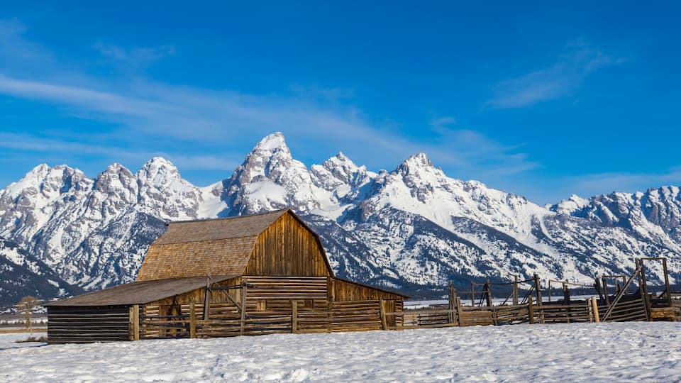A wooden barn in a snowy landscape with snow-capped mountains in the background.