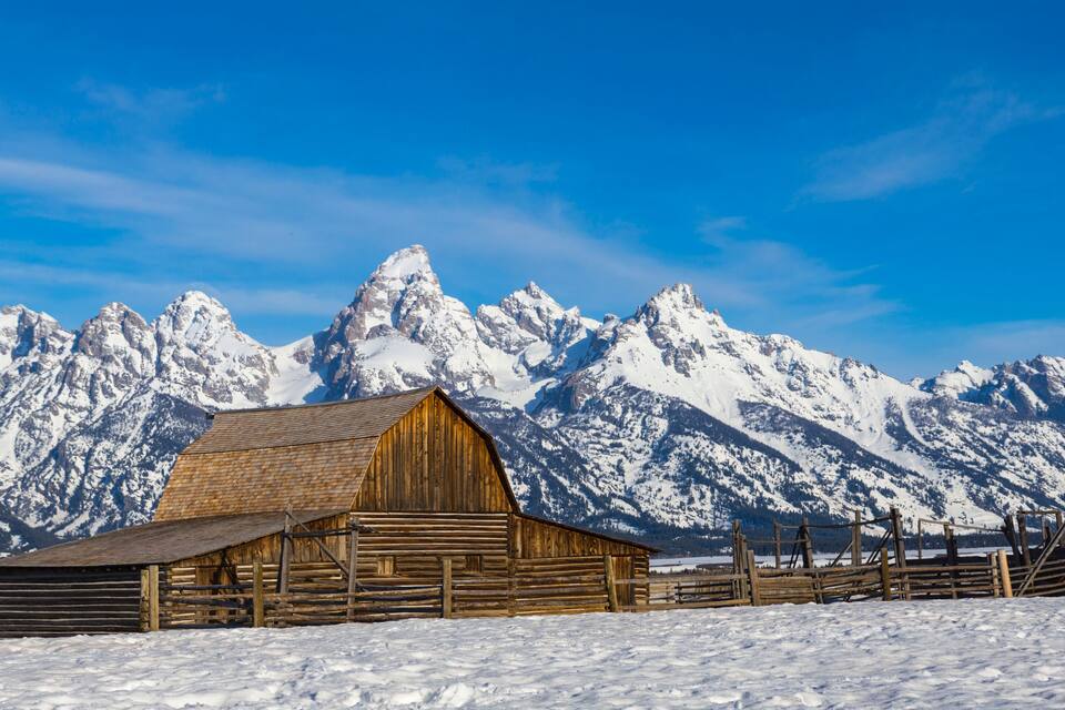 Une grange en bois se dresse dans un paysage enneigé, avec des montagnes aux sommets couverts de neige en arrière-plan.