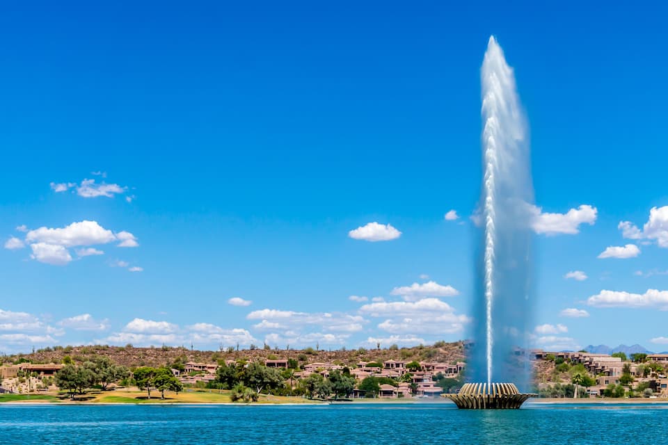 A tall water fountain in a lake with a clear blue sky and a residential area in the background.