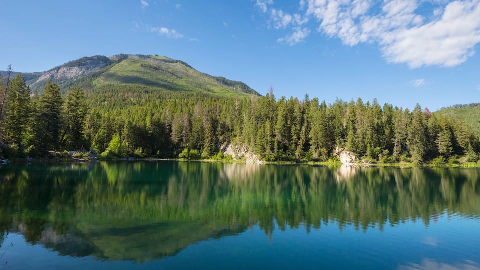 A calm lake with clear reflections, surrounded by dense green forests and a prominent mountain in the background.