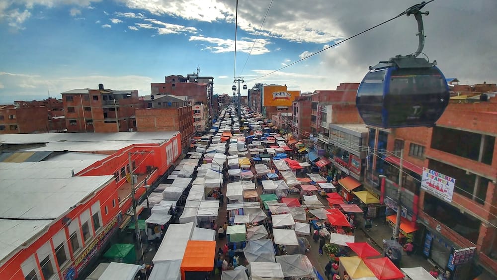 A bustling street market with numerous stalls under colorful umbrellas, set against a backdrop of buildings and a cable car.