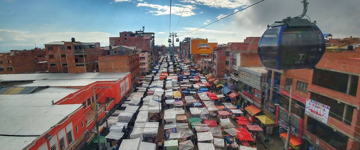 Een levendige straatmarkt met talloze kraampjes onder kleurrijke parasols, met op de achtergrond gebouwen en een kabelbaan.