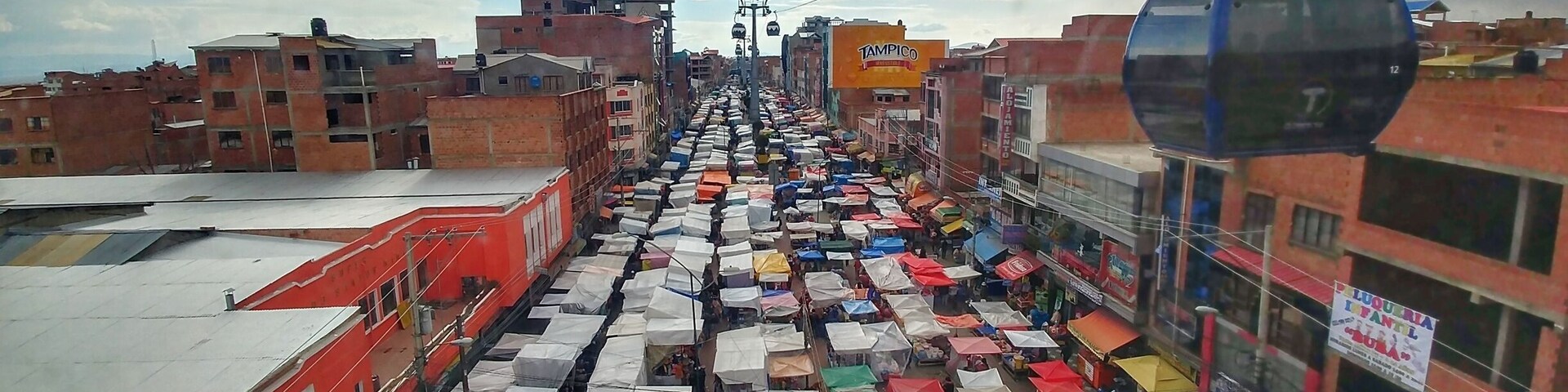 Un mercado callejero concurrido, con numerosos puestos bajo coloridos paraguas, frente a edificios y un teleférico.