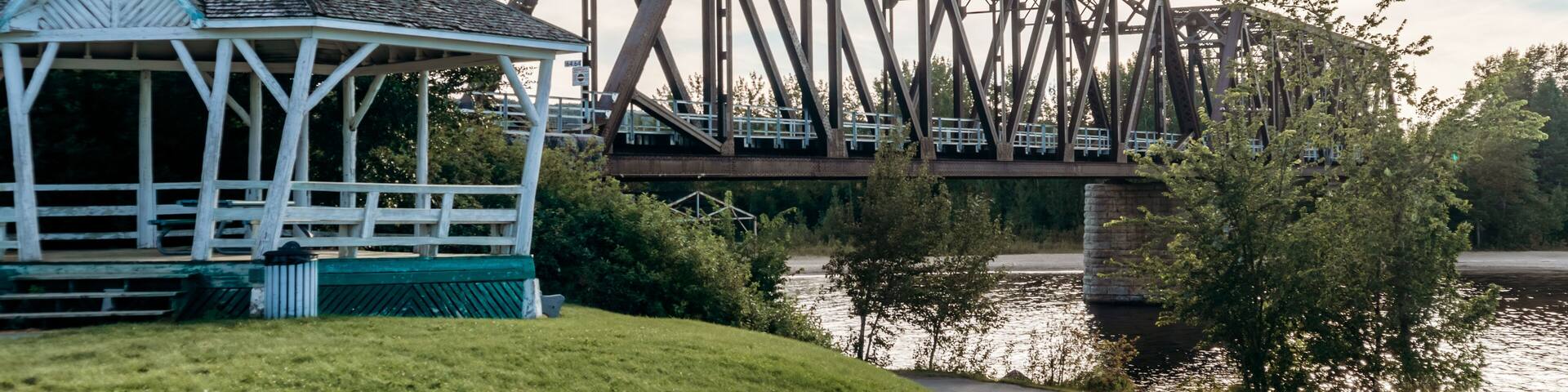 Steel truss bridge crossing a river near the town of Desbiens in Quebec, with riverside park and gazebo in the foreground