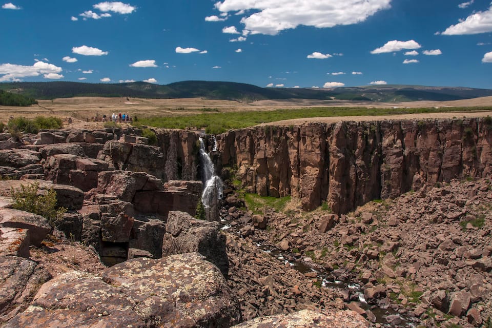 Una cascada se precipita por un risco rocoso, rodeada de un extenso paisaje con colinas lejanas y un cielo azul despejado.
