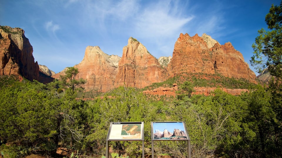A mountainous landscape with prominent red rock formations and dense green vegetation. There are two informational signs in the foreground.