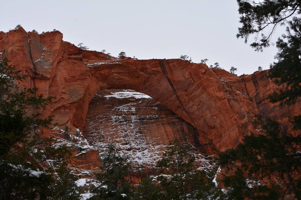 A red rock arch with snow patches and trees in the foreground.