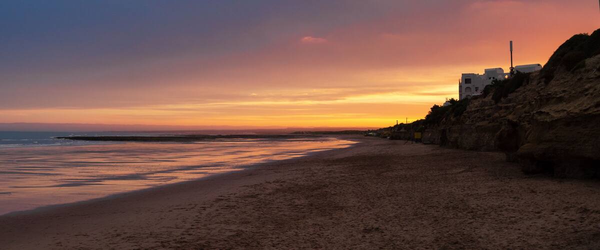 A beach with a sunset, rocky cliffs, and buildings along the coastline.