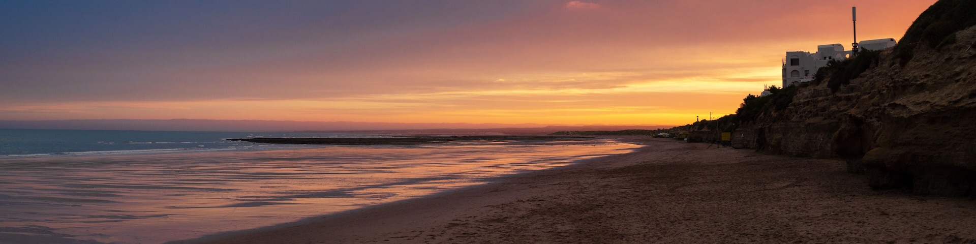 A beach with a sunset, rocky cliffs, and buildings along the coastline.