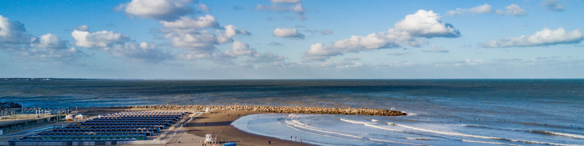 Una playa con un rompeolas rocoso, gente caminando y una hilera de sombrillas.