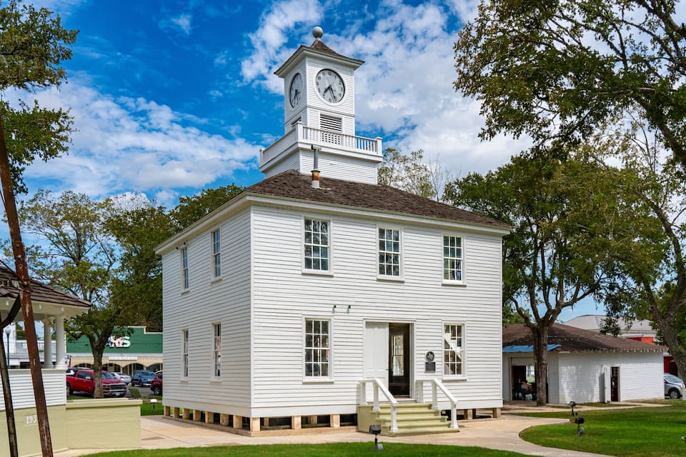 A white two-story building with a clock tower, surrounded by trees and a clear blue sky.