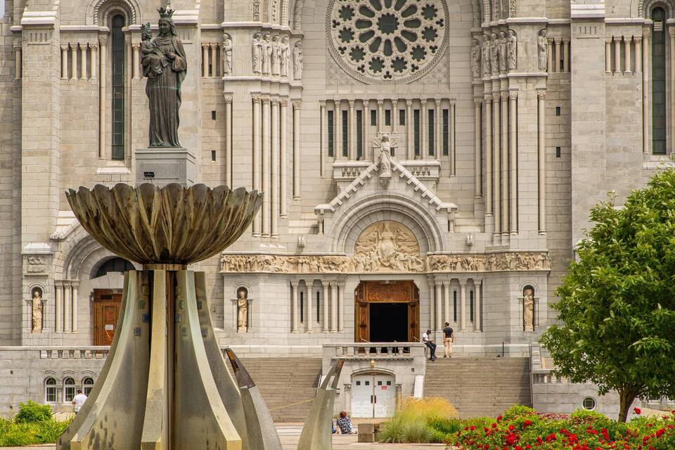 A large stone building with intricate carvings and statues, a central fountain, and a tree with red flowers in the foreground.