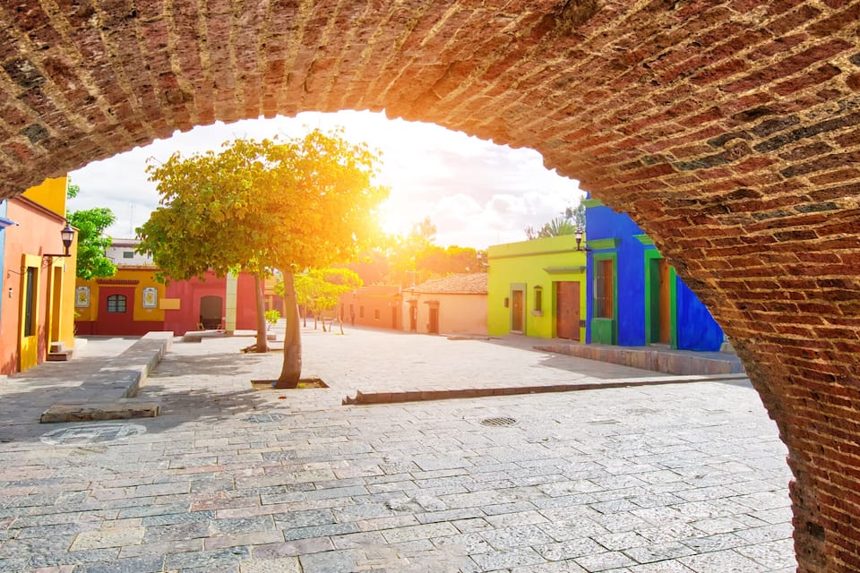 A cobblestone street with colorful buildings, a tree, and a brick archway.