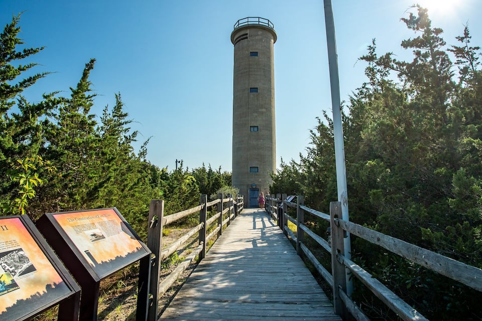 Una passerella di legno che conduce a un'alta torre in cemento con ringhiera metallica, circondata da alberi verdi e fitti.