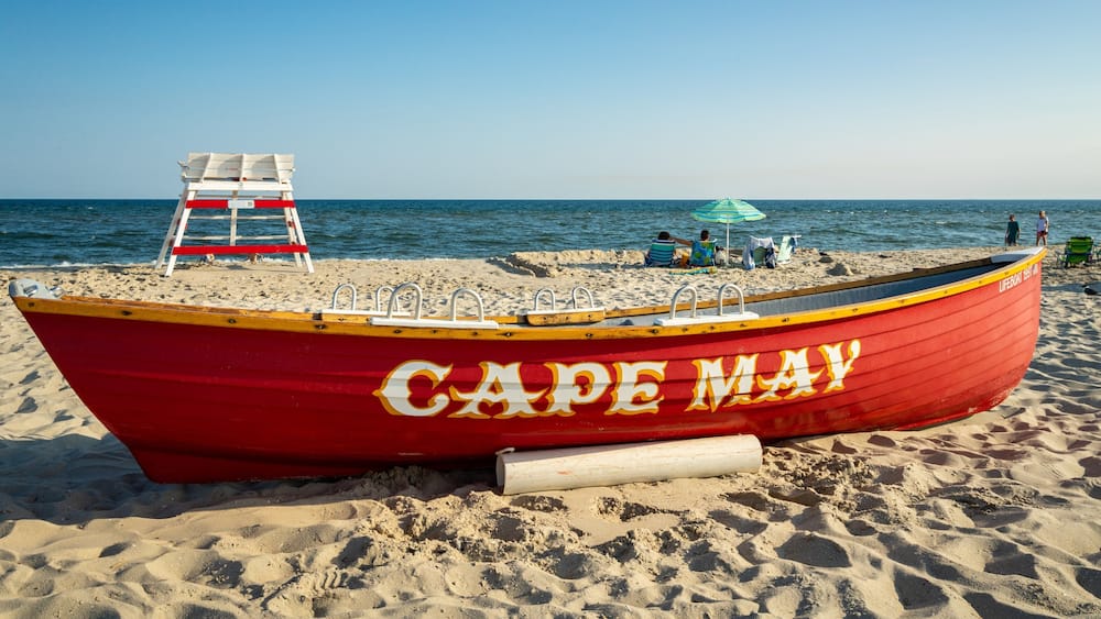 A red lifeboat named "Cape May" on a sandy beach with a lifeguard tower and people in the background.