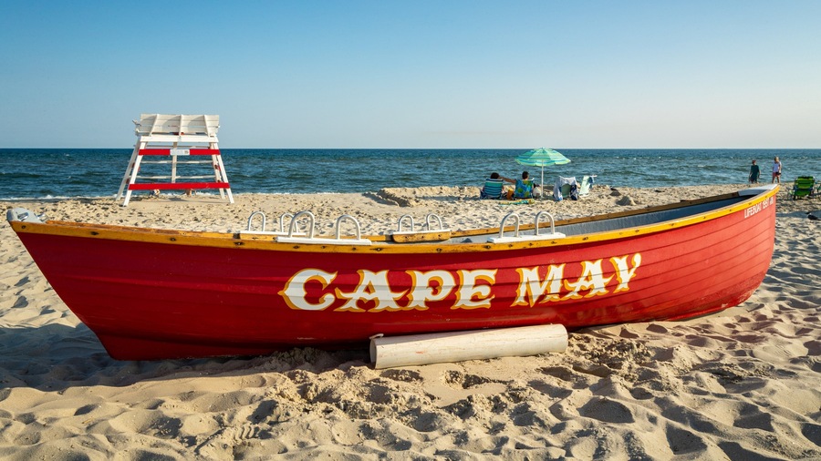 A red lifeboat named "Cape May" on a sandy beach with a lifeguard tower and people in the background.