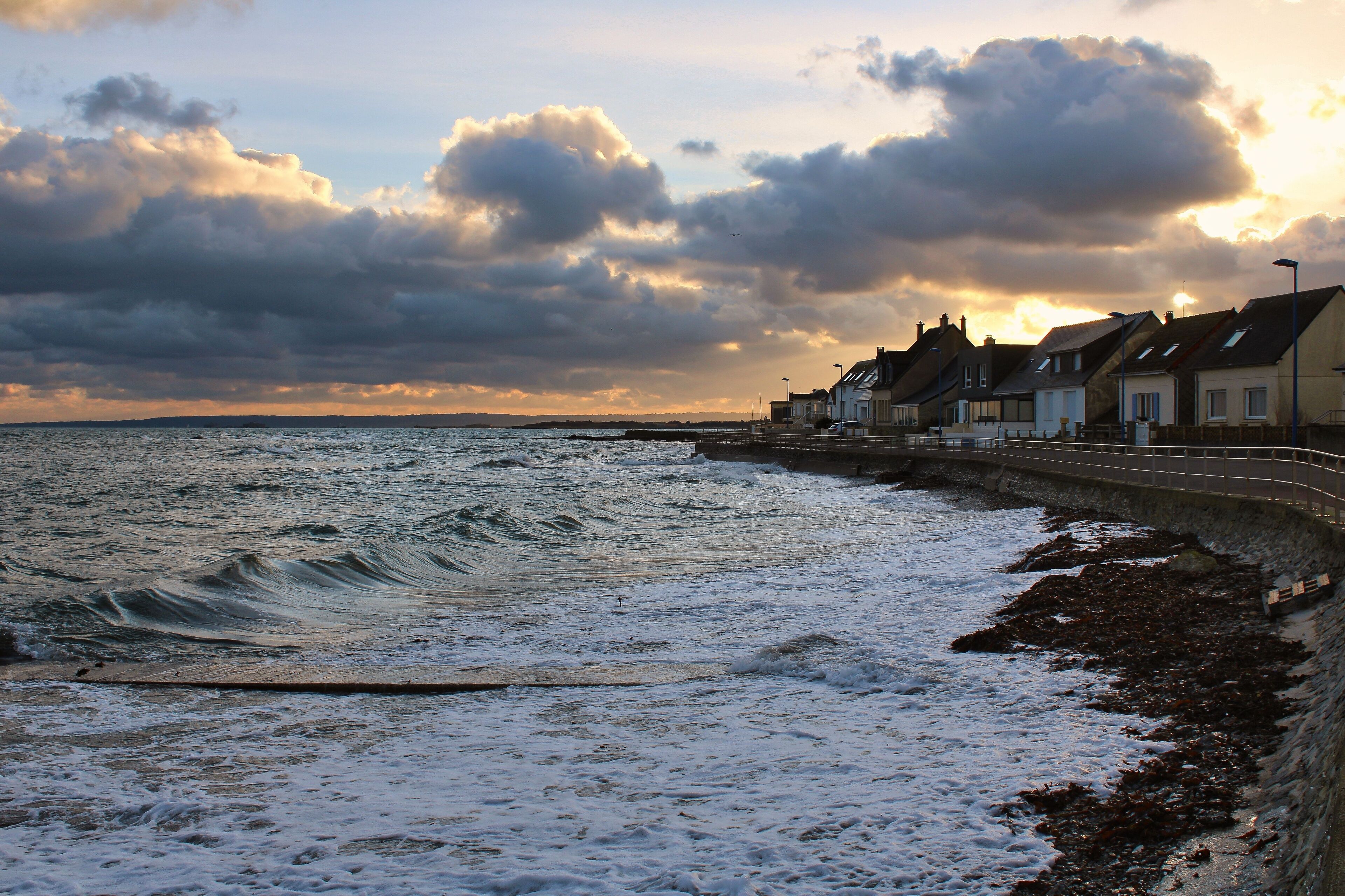 A coastal town with houses along the sea, waves crashing onto the shore, and a cloudy sky.