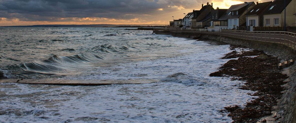A coastal town with houses along the sea, waves crashing onto the shore, and a cloudy sky.