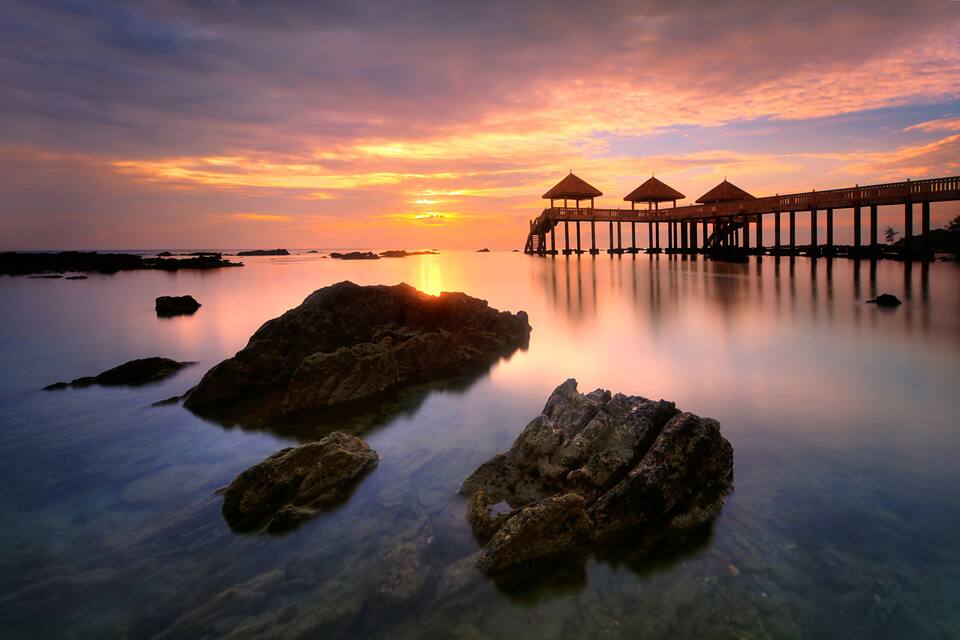 A sunset over a calm sea with a pier and thatched-roof structures.